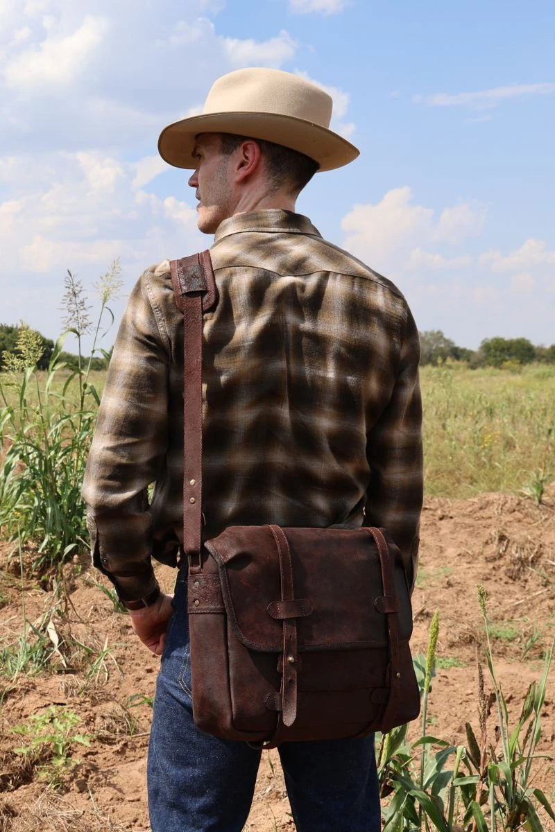 Man wearing a plaid shirt, hat, and carrying a dark brown waxy chocolate boar leather messenger bag in an open field damaged by boar