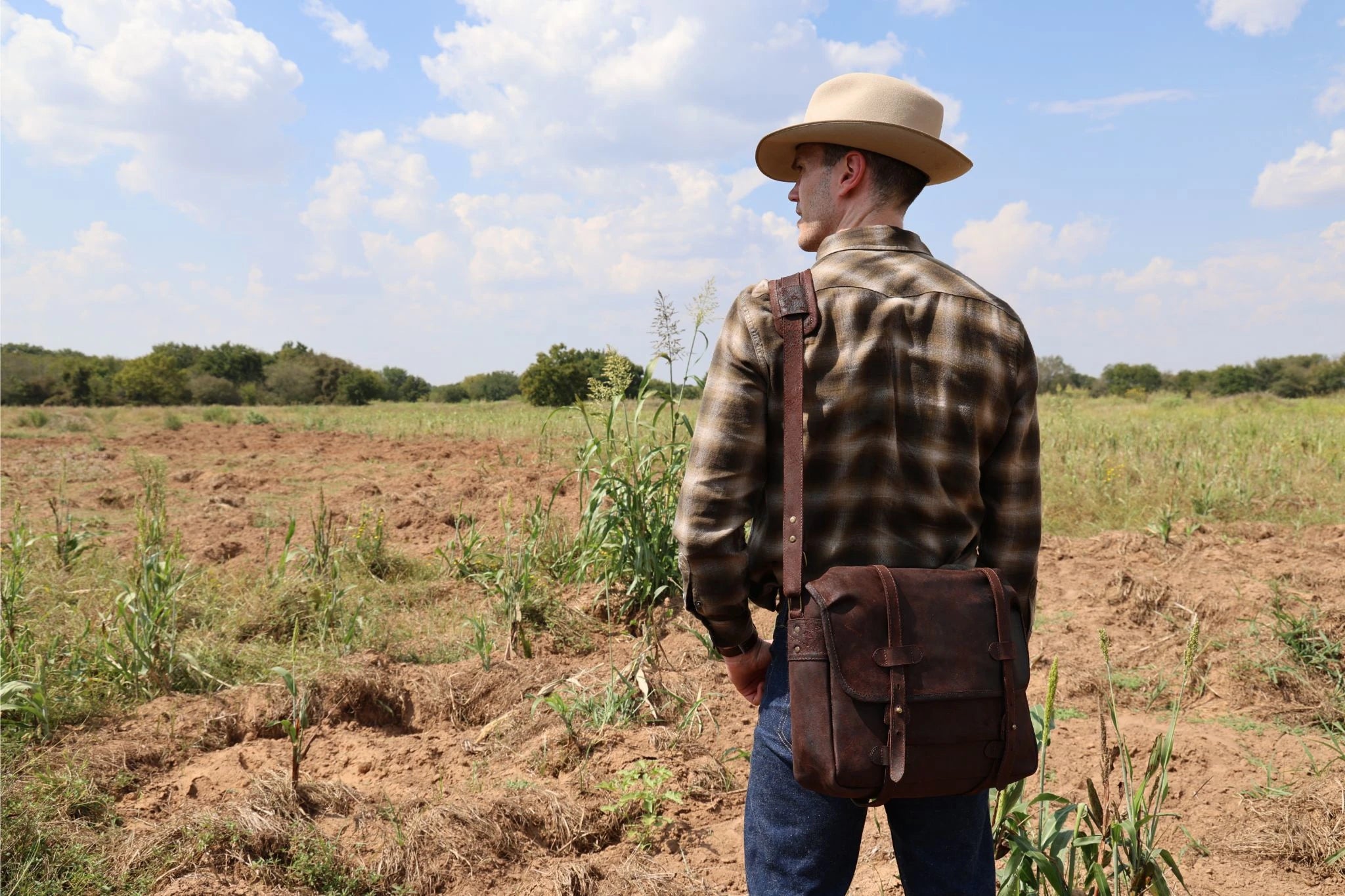 Man in a plaid shirt and hat standing in a field with a leather bag over his shoulder.