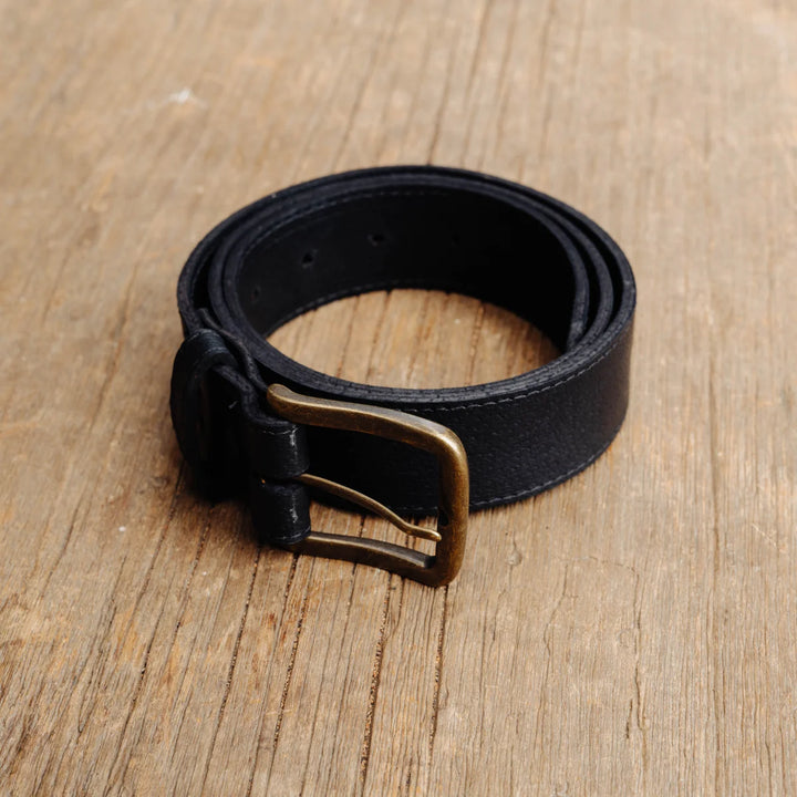 A black feral hog leather belt with a brass buckle, coiled on a light brown wooden surface.