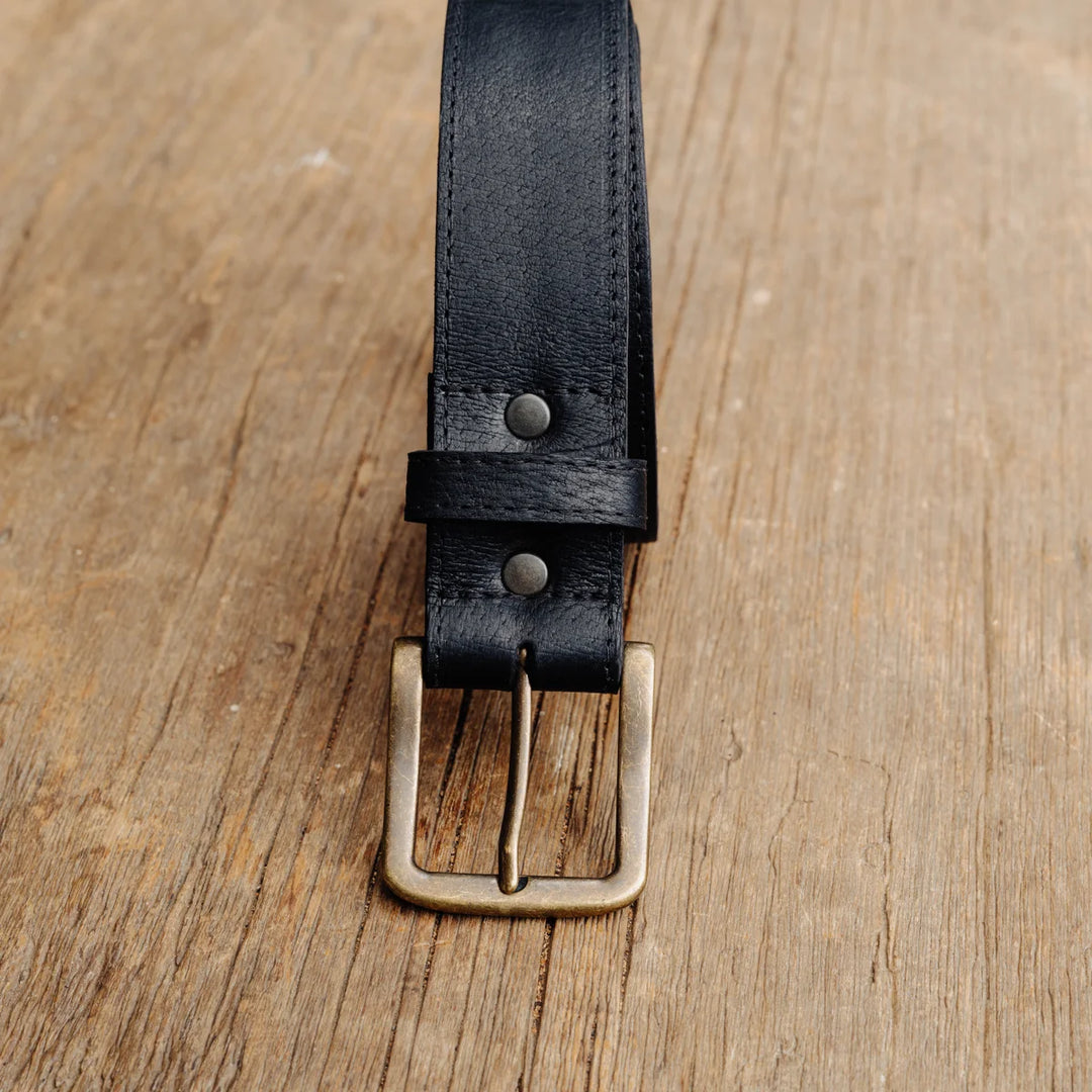 A close-up of a black feral hog leather belt with a brass buckle, laid flat on a rustic wooden surface.