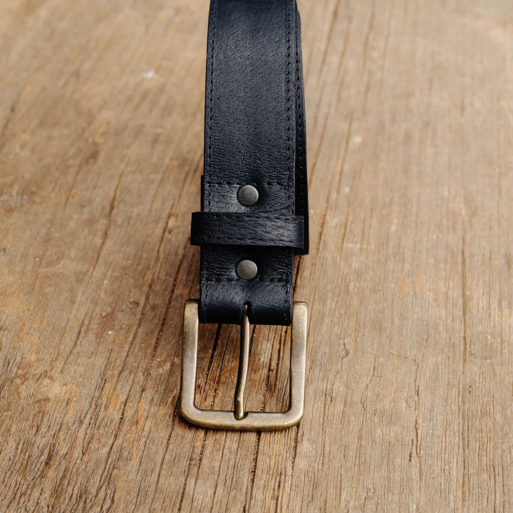 A close-up of a black feral hog leather belt with a brass buckle, laid flat on a rustic wooden surface.
