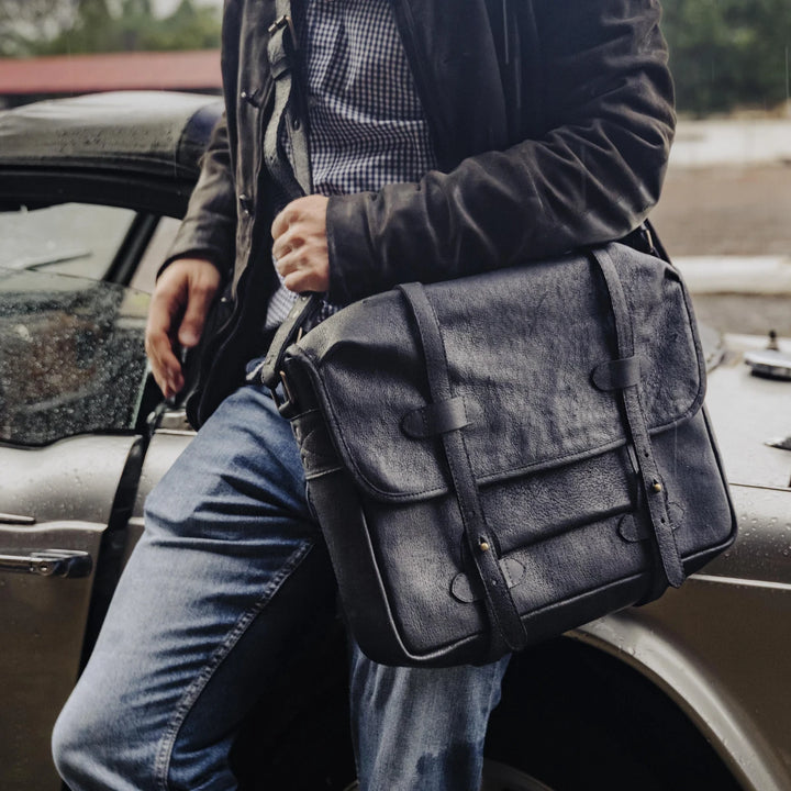 Man holding a black boar leather messenger bag next to a vintage car.