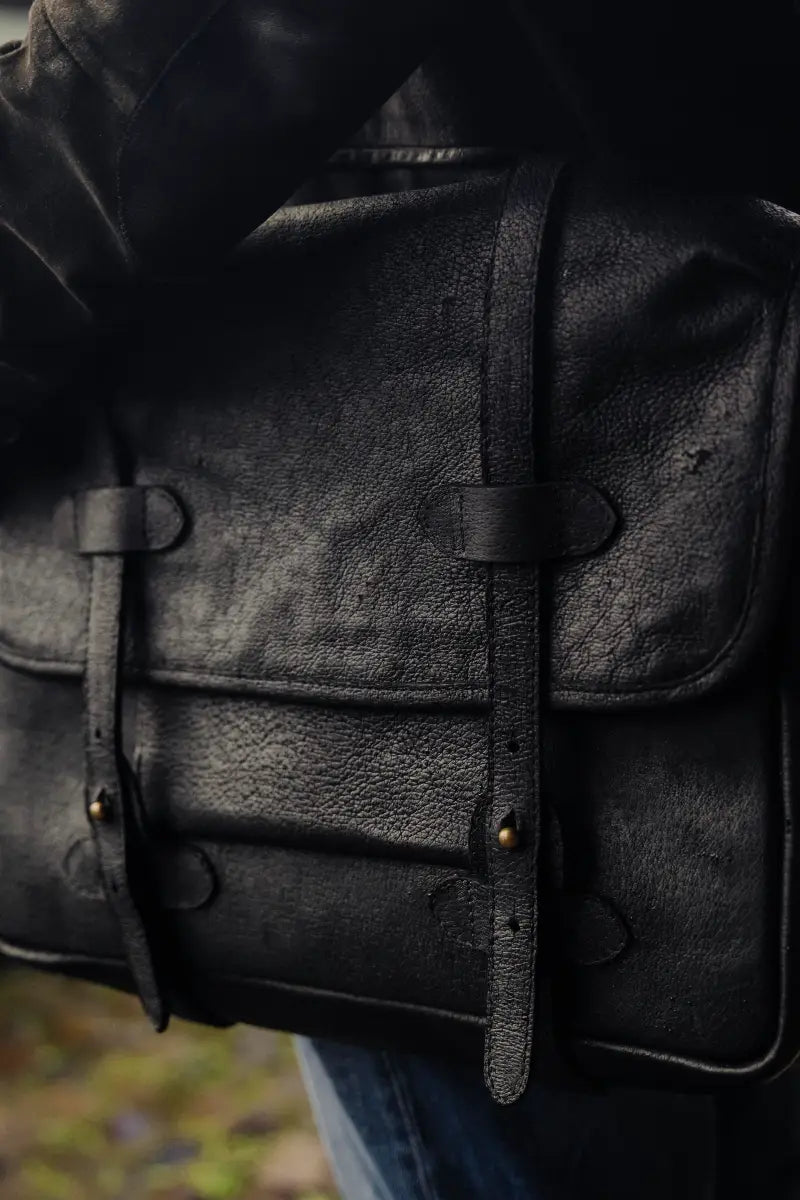Dark boar leather bag with visible stitching and leather straps on a blurred background