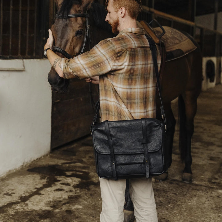 Man with a plaid shirt and black jabari boar leather bag standing next to a horse in a stable.