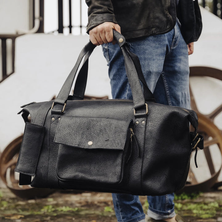 Man holding a black jabari boar leather duffel bag outdoors.
