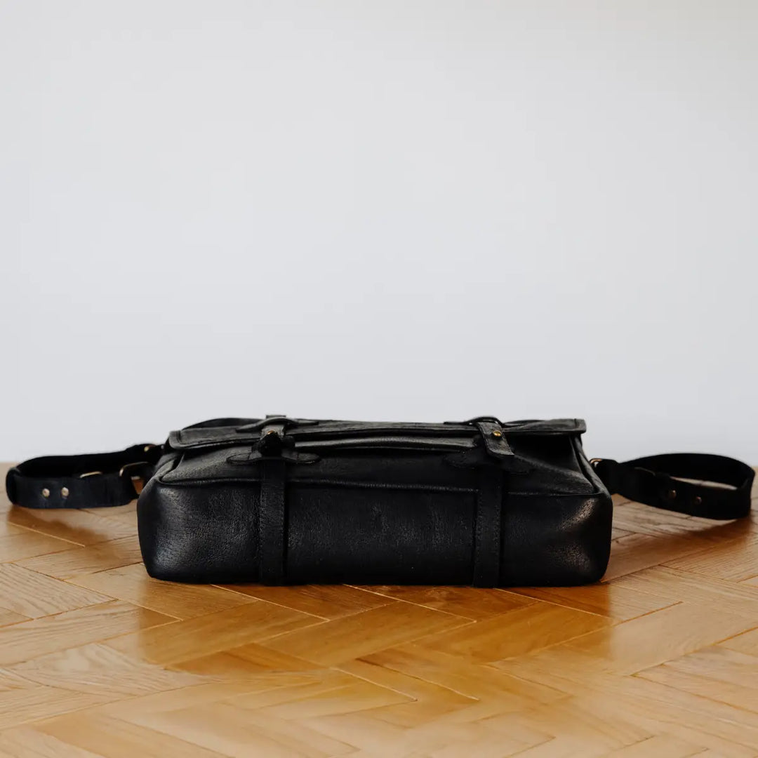 Black boar leather messenger bag lyaing flat on a wooden floor with a light gray background