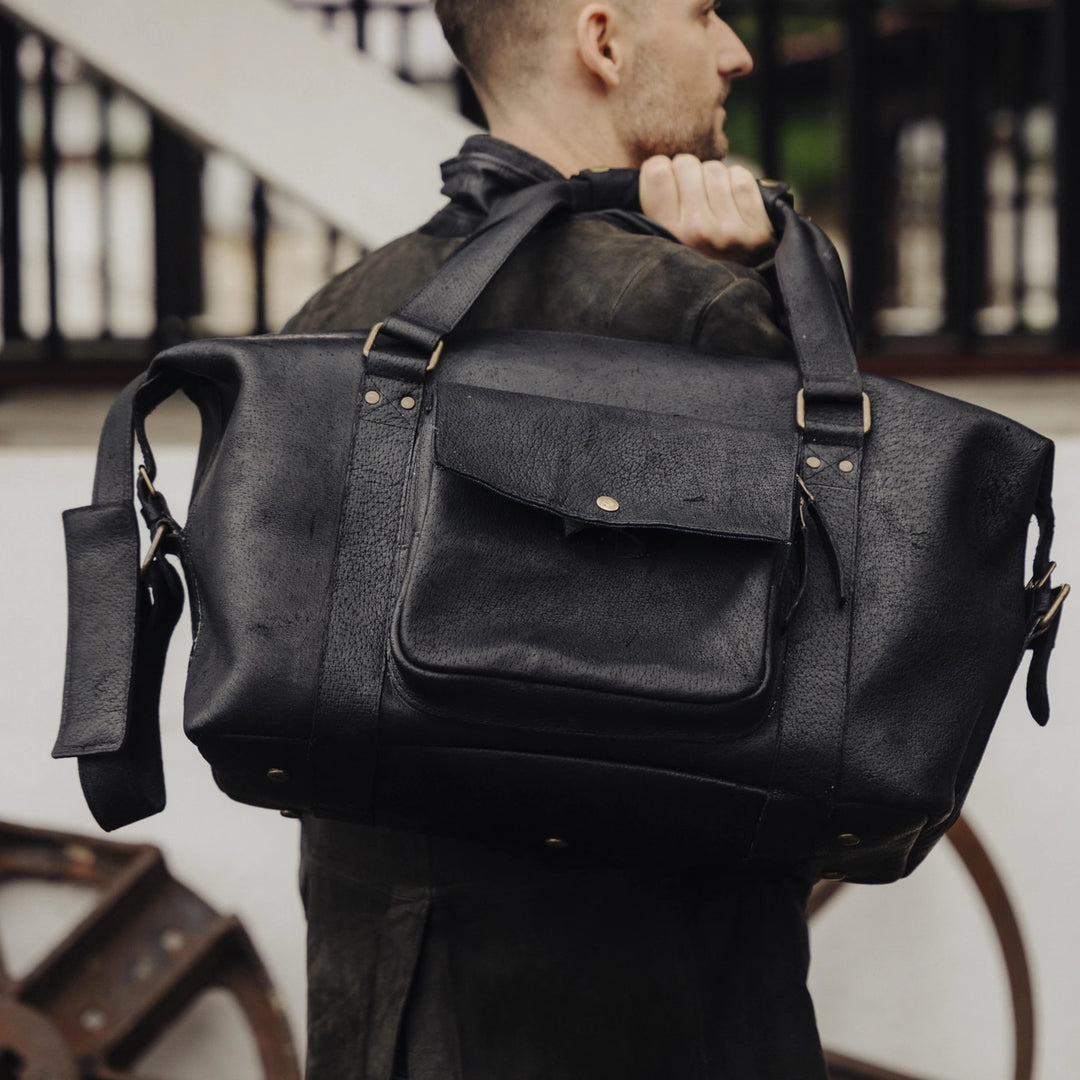A man carrying a large black boar leather duffel bag in an outdoor setting.