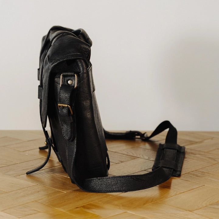 Black leather messenger bag on a wooden floor with a plain background