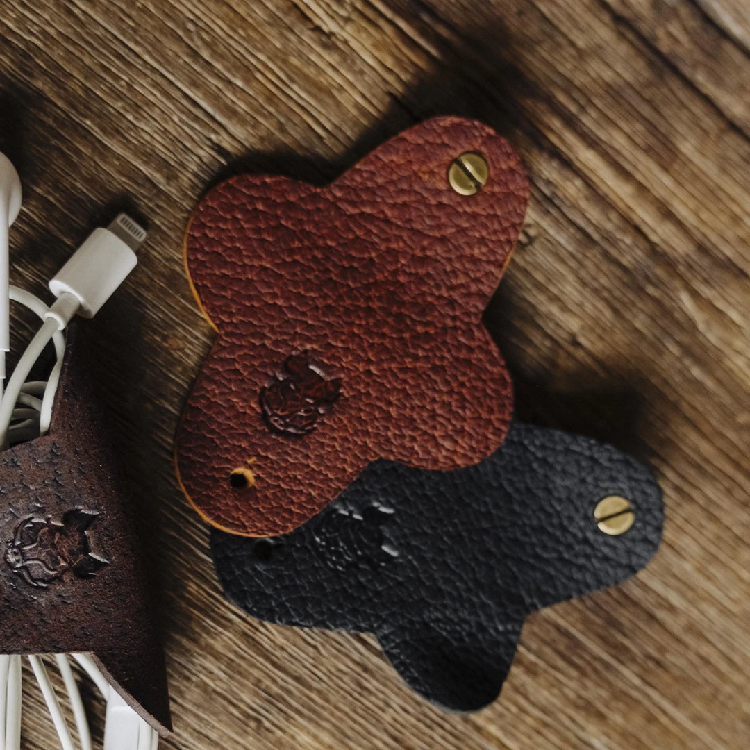 Two boar leather cable wraps, one brown and one black, on a wooden surface with a USB cable.
