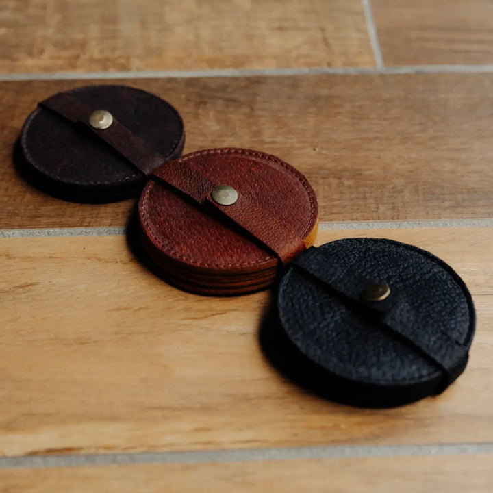 Three leather coasters in black, brown, and dark brown on a wooden surface.