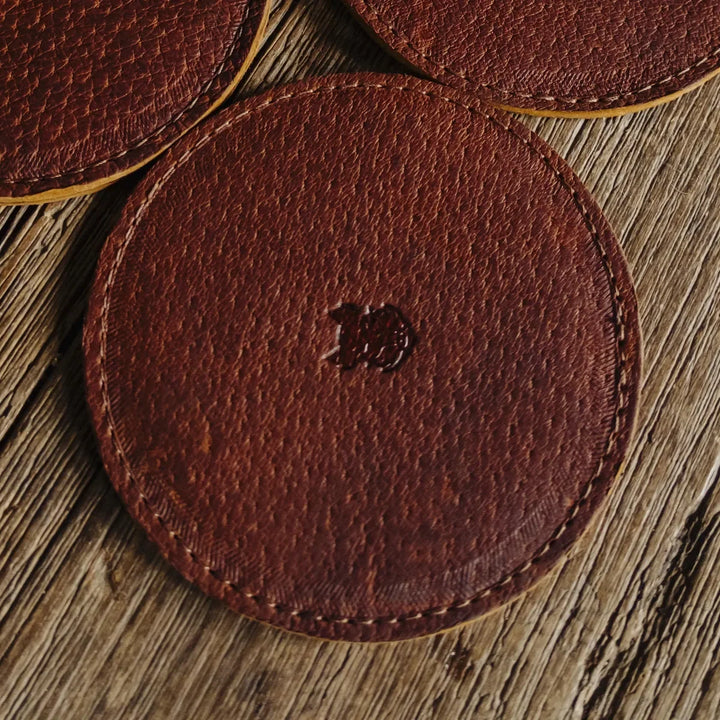 Brown leather coasters with a Bad Leather Goods logo on a wooden surface