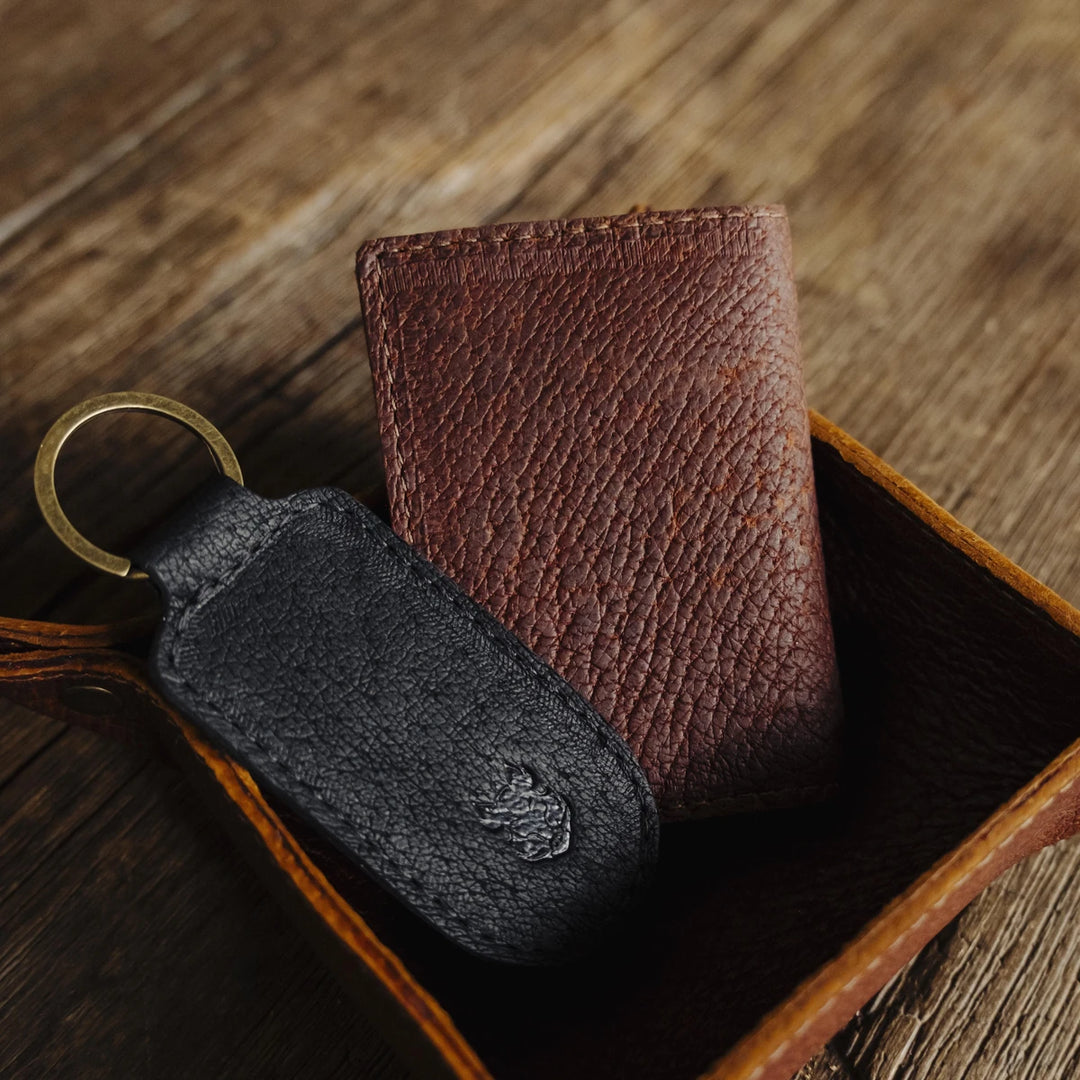 One brown boar leather keychain and one black boar leather wallet, in a brown boar leather valet tray, on a wooden surface.