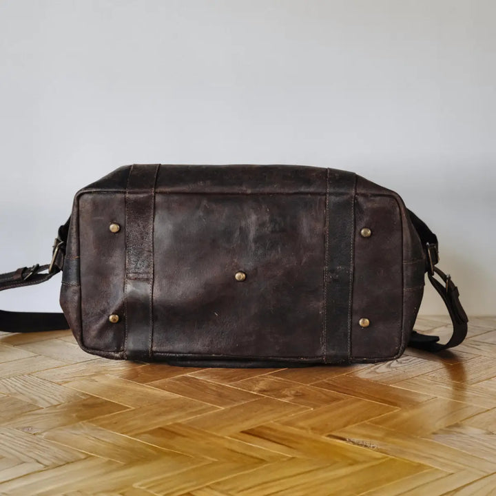 Dark brown boar leather duffel bag on a wooden floor with a white wall background
