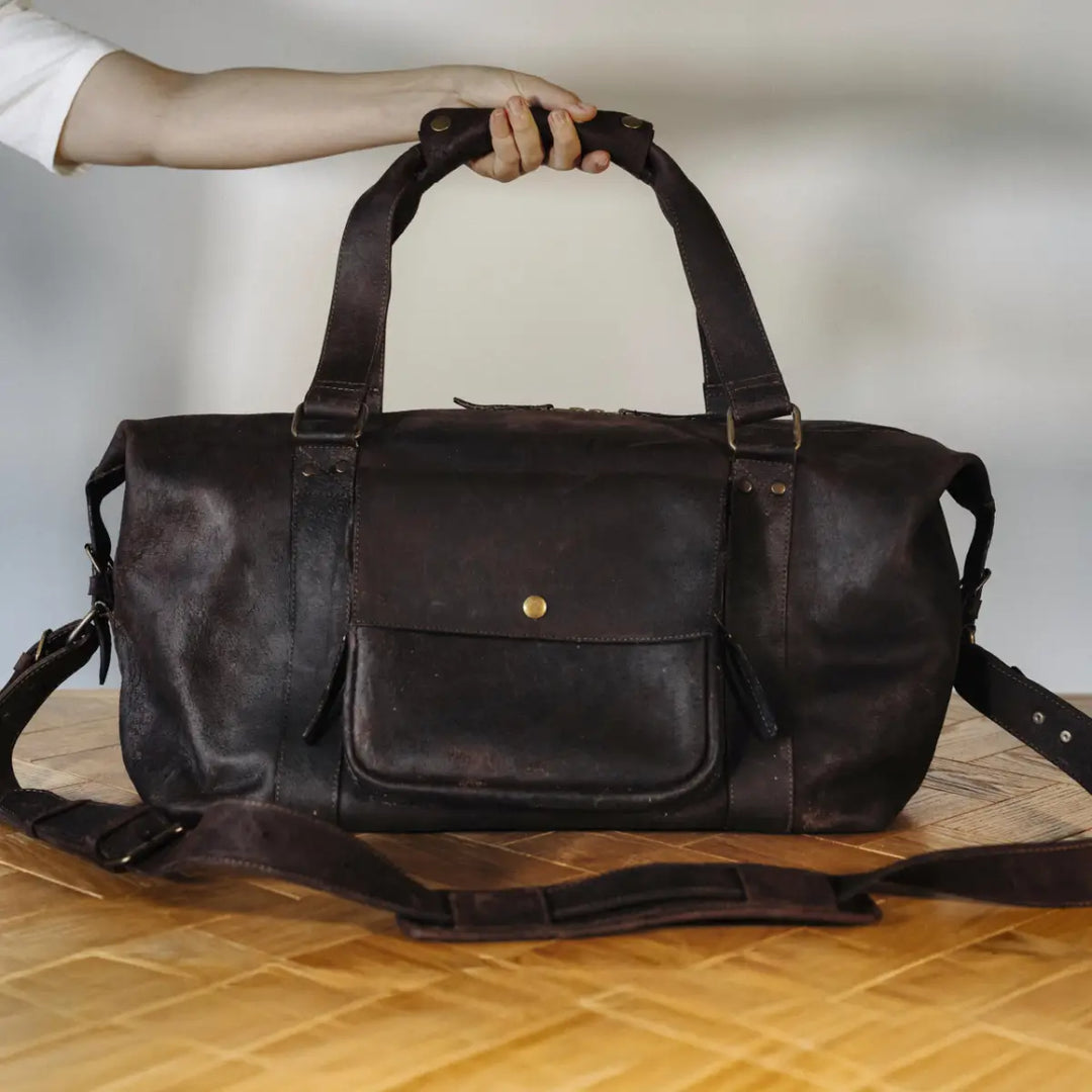 Brown waxy chocolate boar leather duffel bag held by a hand on a wooden surface with a neutral background