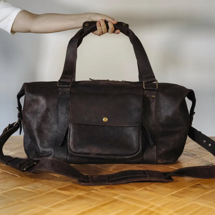 Brown waxy chocolate boar leather duffel bag held by a hand on a wooden surface with a neutral background