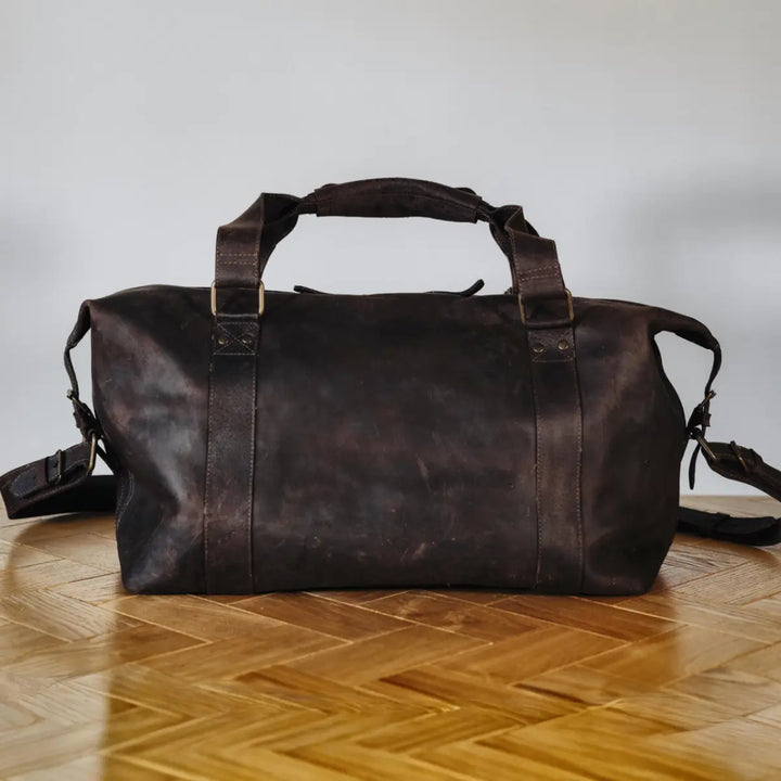 Dark brown boar leather duffel bag on a wooden floor with a neutral background