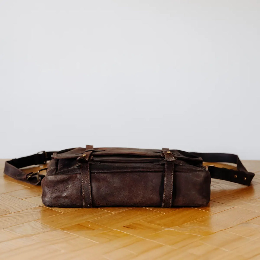 Brown boar leather messenger bag lyaing flat on a wooden floor with a light gray wall background