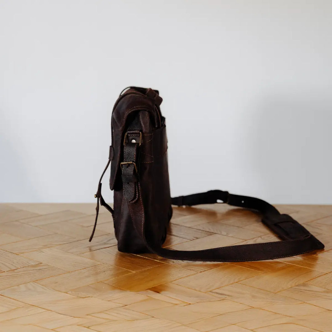 Dark brown boar leather messenger bag side view on a wooden floor with a white wall background