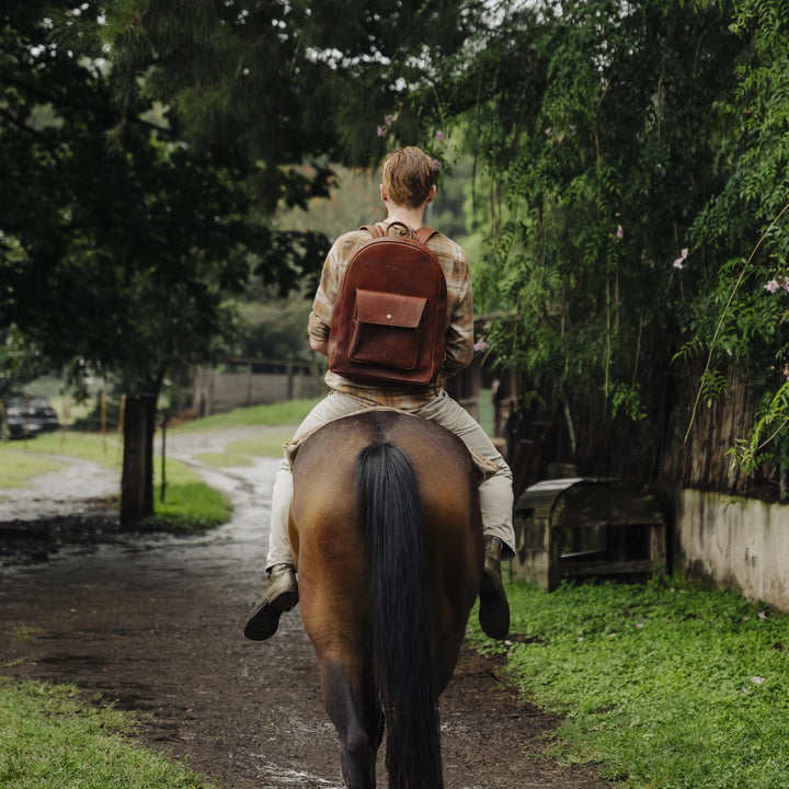 Person riding a horse on a path surrounded by greenery wearing a brown boar leather backpack