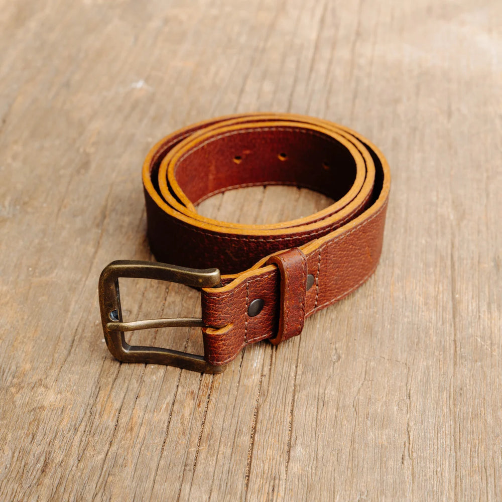 A cognac brown feral hog leather belt with a metal buckle, coiled on a rustic wooden surface.