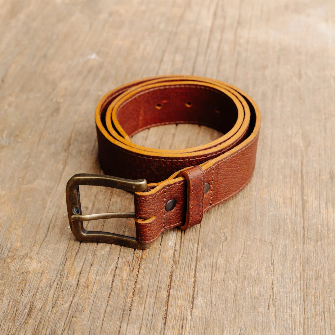 A cognac brown feral hog leather belt with a metal buckle, coiled on a rustic wooden surface.