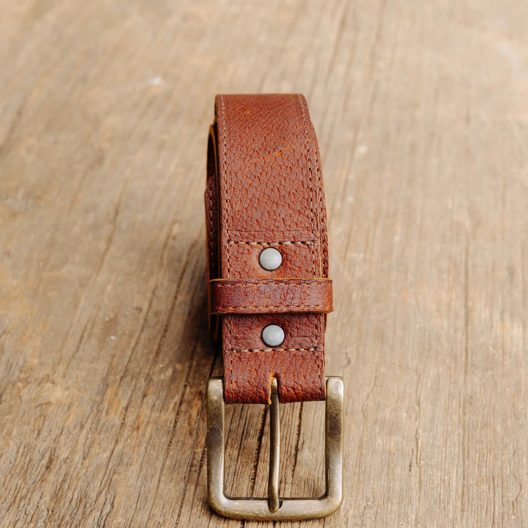 A cognac brown textured feral hog leather belt with a brass buckle, laid flat on a rustic wooden surface.