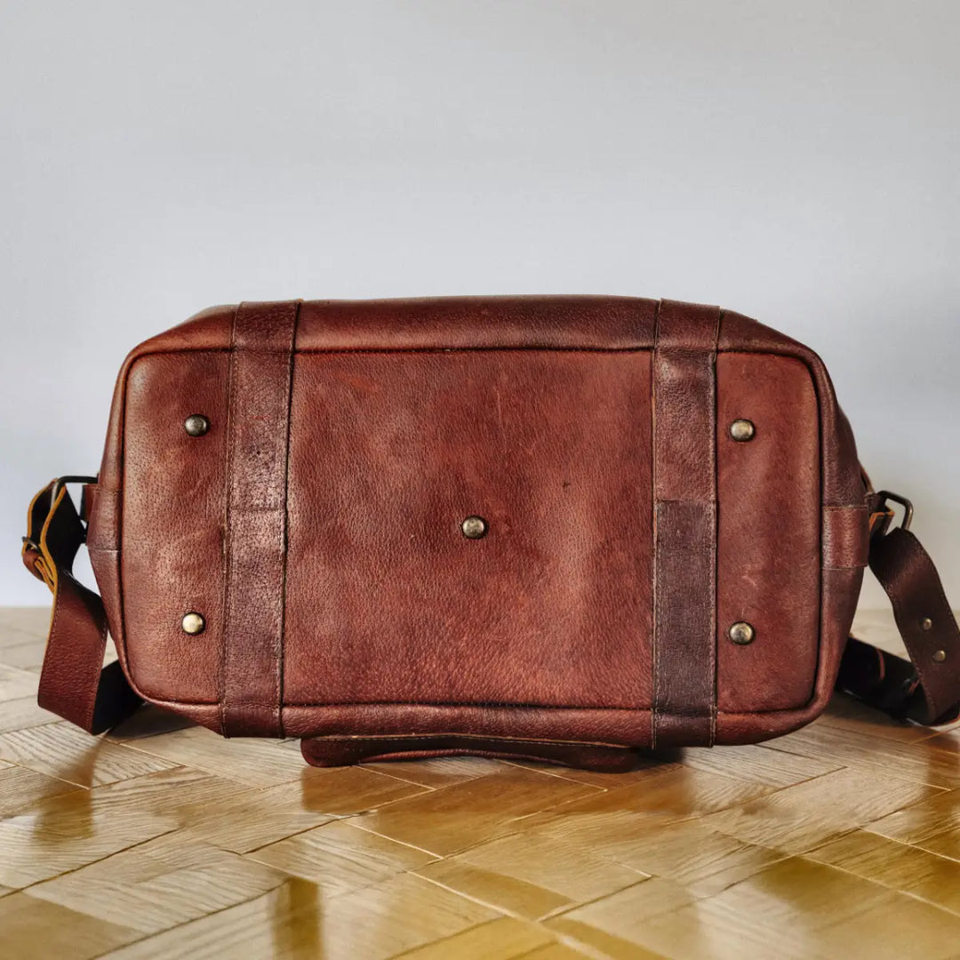 The bottom of a brown boar leather bag with metal studs on a wooden floor with a light gray wall background