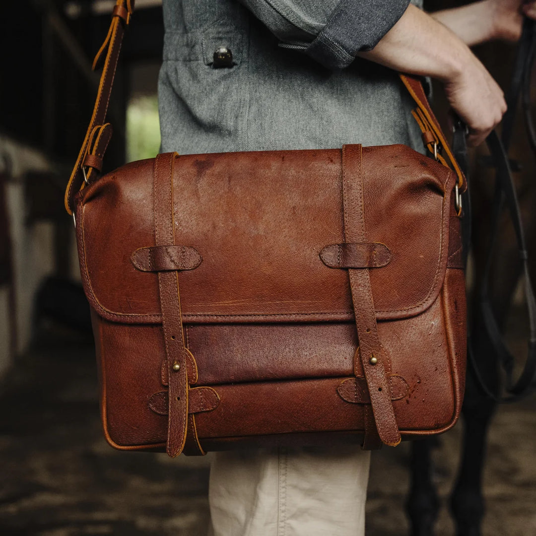 Brown cognac jabari boar leather messenger bag held by a man in a barn petting a horse