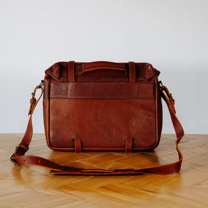 Brown boar leather messenger bag, with visible luggage strap, on a wooden floor with a light gray wall background