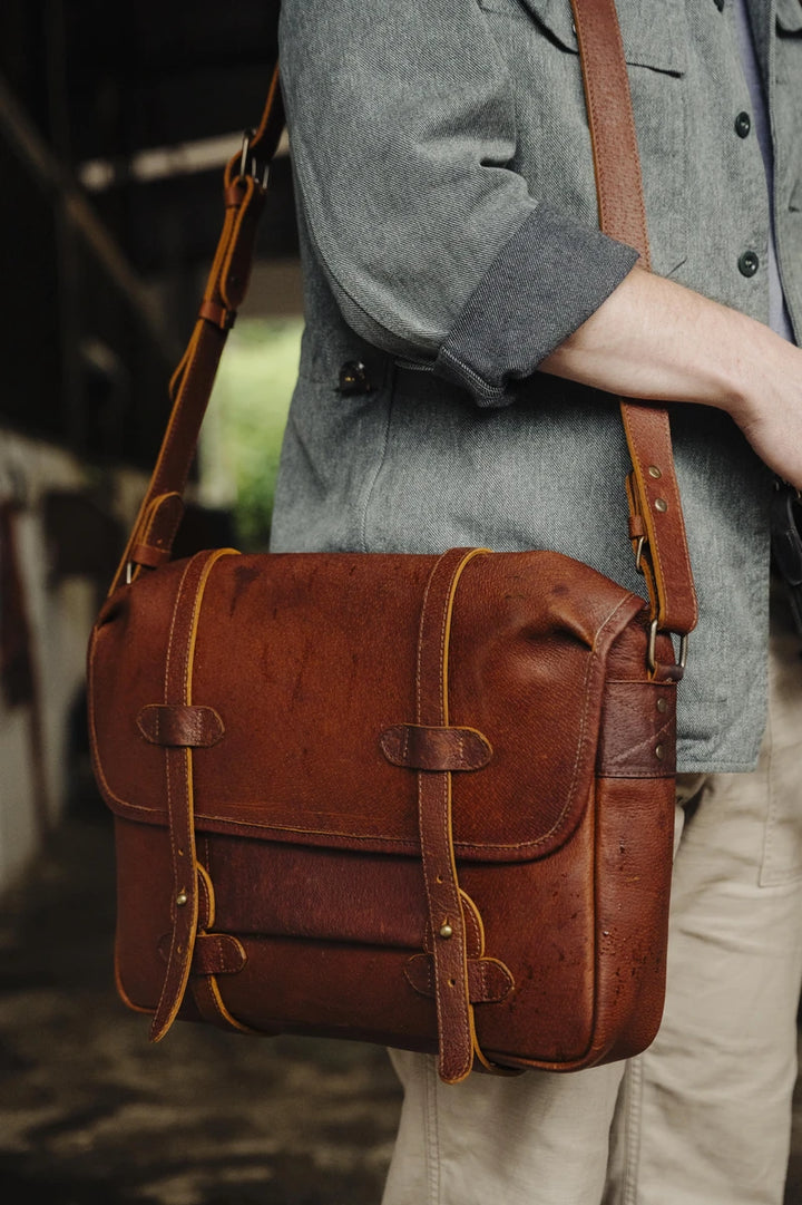 Brown cognac leather messenger bag worn by a man in a gray coat.