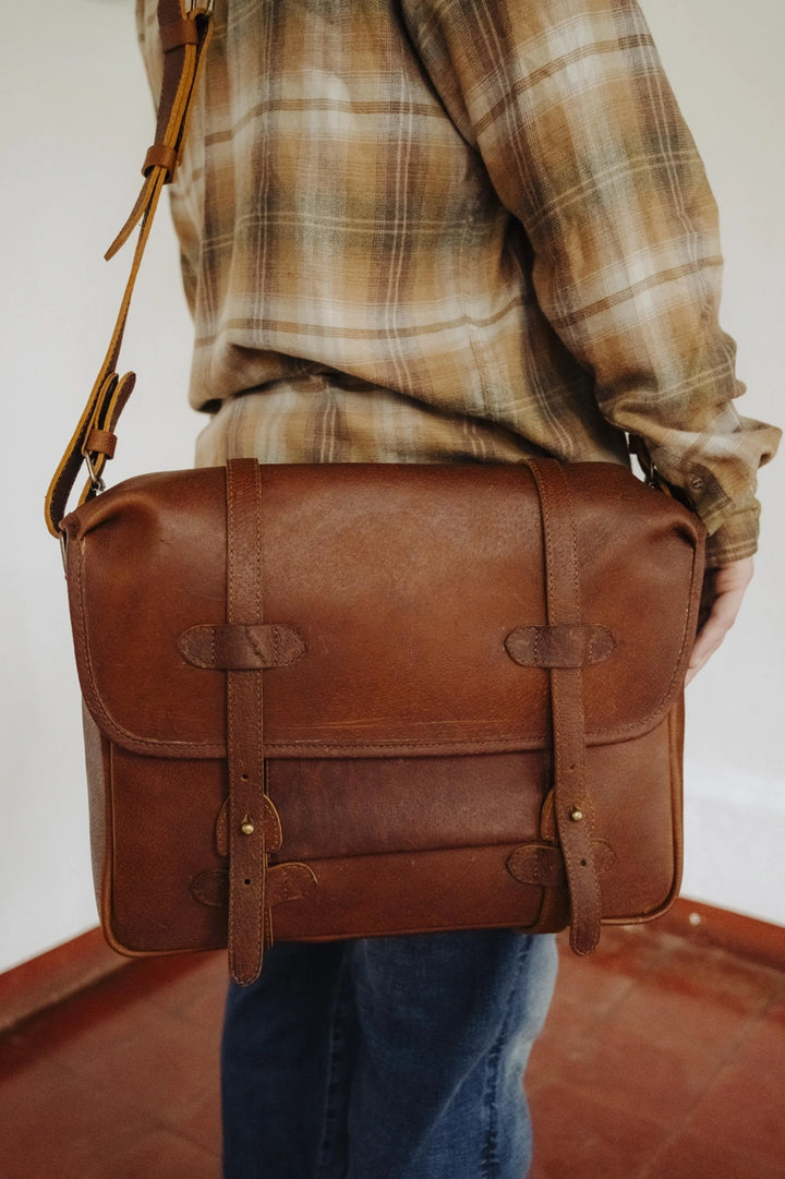 Man holding a brown boar leather messenger bag with a plaid shirt and jeans.