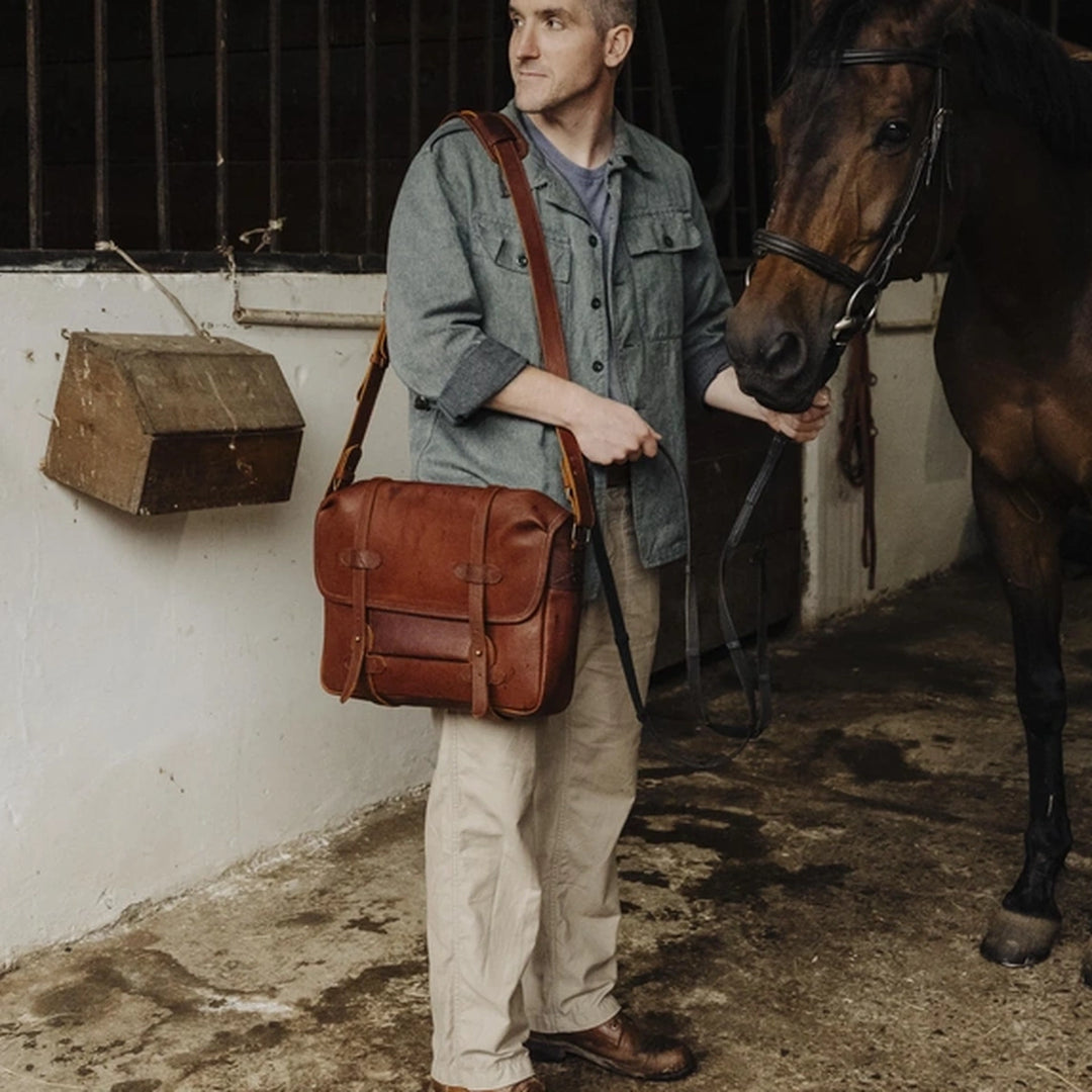 A man holding a brown cognac Jabari leather messenger bag next to a horse in a stable.