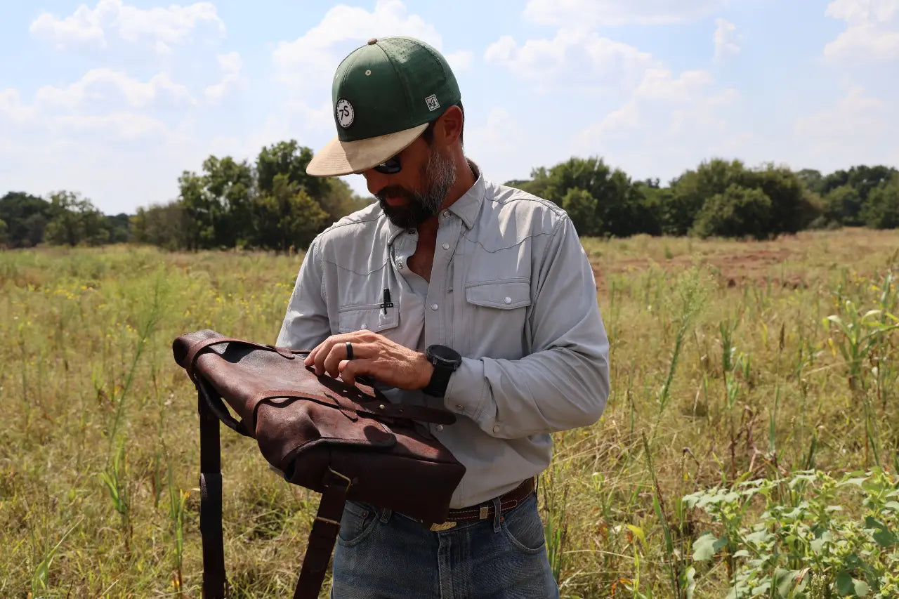 Man holding a leather bag in a field with trees and blue sky in the background