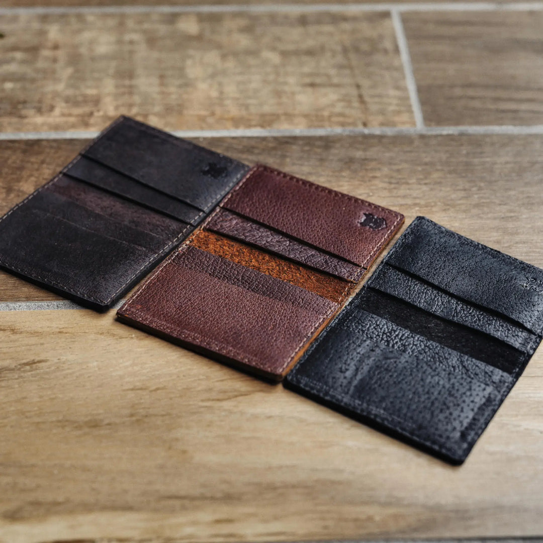 Three boar leather card wallets in black, brown, and dark brown on a wooden surface.