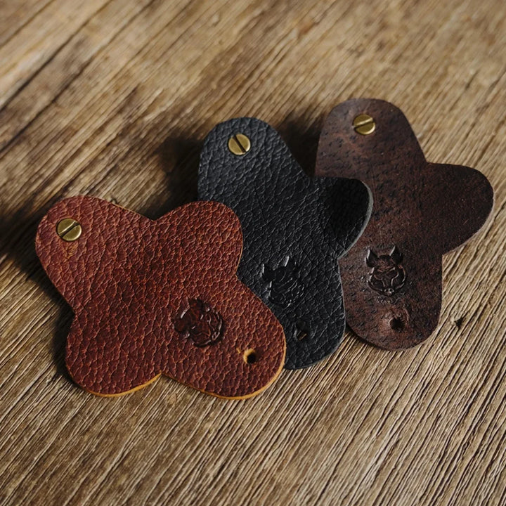 Three boar leather cable wraps in brown cognac jabari, black jabari, and dark brown chocolate waxy boar on a wooden surface.