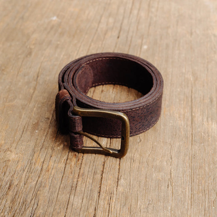 A dark brown feral hog leather belt with a brass buckle, coiled on a light brown wooden surface.