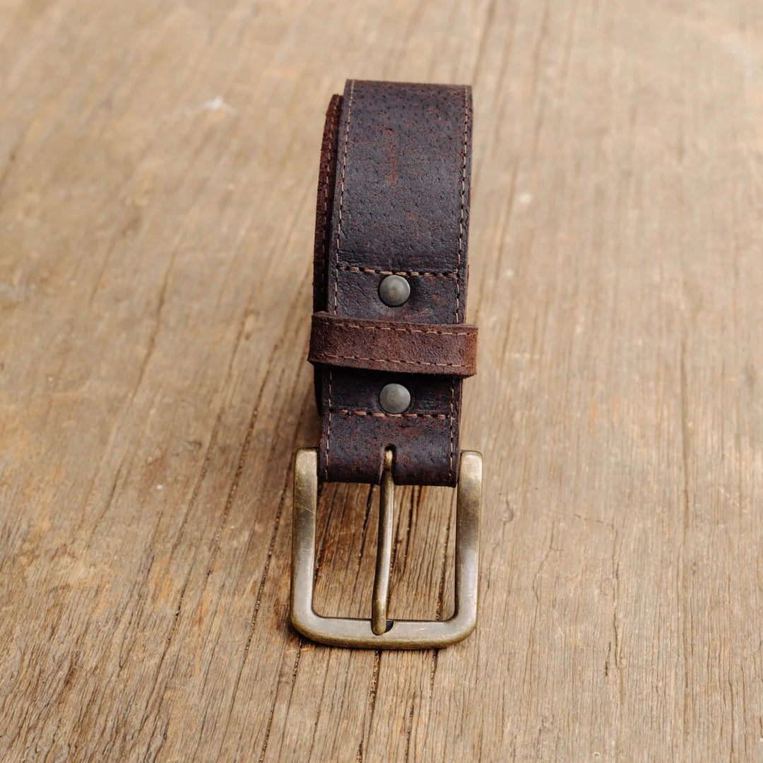 A dark brown feral hog leather belt with a brass buckle, neatly coiled and displayed on a rustic wooden surface.
