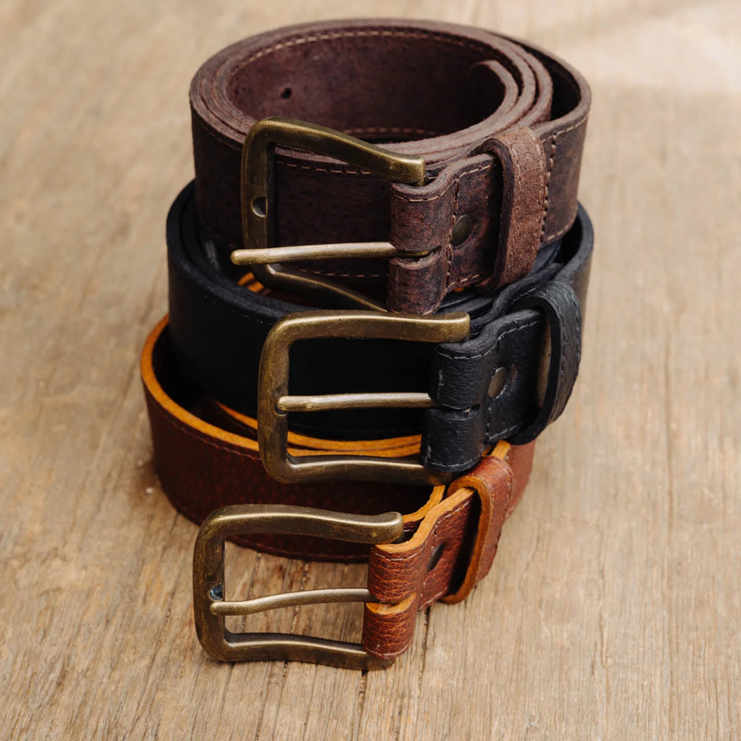 Three feral hog leather belts in brown, black, and cognac stacked on a light wood surface, each with a brass-tone buckle.