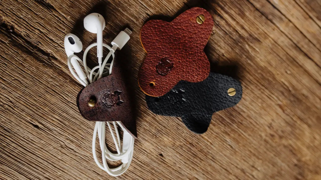 Three leather cable holders in brown, black, and red on a wooden surface with white earbuds.
